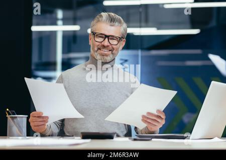 Homme d'affaires prospère avec des documents souriants et regardant l'appareil photo, portrait de l'investisseur à cheveux gris mature financier dans le bureau moderne, homme derrière le papier travail avec rapports comptes et contrats Banque D'Images