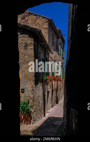 Volets de fenêtre verts, géraniums rouges et ciel bleu clair : une rangée de maisons en ruines de pierre vue à travers une arche à Pienza, un village perché dans le pittoresque Val d'Orcia en Toscane, en Italie, qui a été reconstruite au milieu de 1400s comme un refuge papal de Rome. Banque D'Images