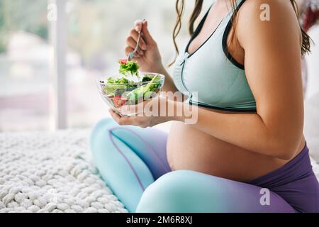 Gros plan portrait d'une femme enceinte en vêtements de sport mangeant une salade fraîche assise au lit Banque D'Images
