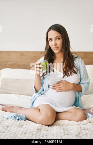 Portrait de la jeune femme enceinte en pajama buvant du jus de smoothie Detox vert assis dans le lit Banque D'Images
