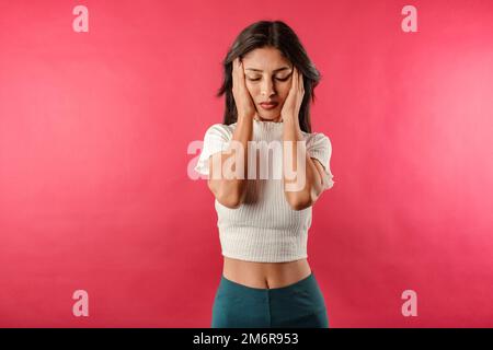 Portrait d'une jeune femme brunette portant une récolte blanche striée isolée sur fond rouge tenant les mains de chaque côté de la tête semble très fatiguée et enfanté Banque D'Images