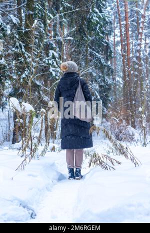 Femme debout dans la forêt d'hiver pendant les vacances de promenade, marcher dans les bois seul avec sac fourre-tout, en bonnet tricoté, veste de puffer. Photo de haute qualité Banque D'Images