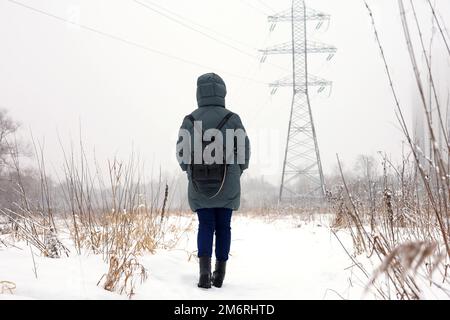 Femme portant une veste en duvet marchant sur le fond des tours électriques par temps neigeux. Loisirs dans le parc d'hiver, saison froide Banque D'Images