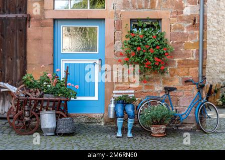 Curieuse décoration sur une porte d'entrée, jeans bleu pantalons en bottes de caoutchouc avec pot de fleur, géraniums, vélo, vieille échelle chariot avec bois de chauffage et lait Banque D'Images