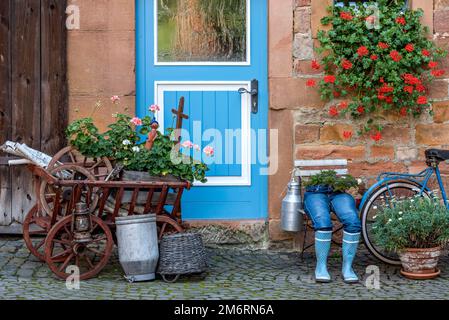 Curieuse décoration sur une porte d'entrée, jeans bleu pantalons en bottes de caoutchouc avec pot de fleur, géraniums, vélo, vieille échelle chariot avec bois de chauffage et lait Banque D'Images