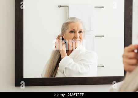 Portrait d'une femme âgée avec un peigne de bois regardant un miroir. Femme âgée peignant ses beaux cheveux longs gris. Banque D'Images