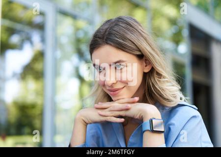 Portrait en gros plan de femme blonde souriante, étudiante avec montre numérique, regardant l'appareil photo, assis dans la rue Banque D'Images