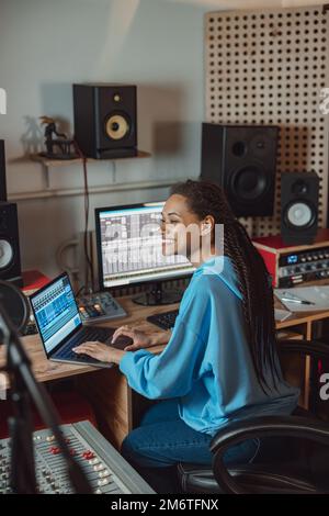 Jeune femme africaine souriante ingénieur du son, compositeur travaille sur ordinateur portable dans le bureau de présentateur radio du studio d'enregistrement audio Banque D'Images