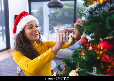 Bonne femme afro-américaine portant le chapeau du père noël, décorant l'arbre de noël dans la salle de séjour Banque D'Images