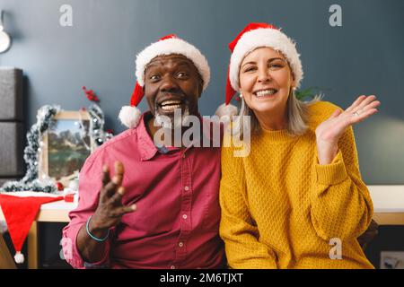 Joyeux couple senior varié à santa chapeaux pour les appels vidéo de noël, souriant et faisant des gestes à la caméra Banque D'Images