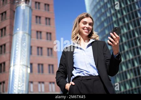 Prise de vue en contre-plongée d'une femme d'affaires en costume, debout dans la rue et regardant un téléphone portable, tenant un smartphone Banque D'Images