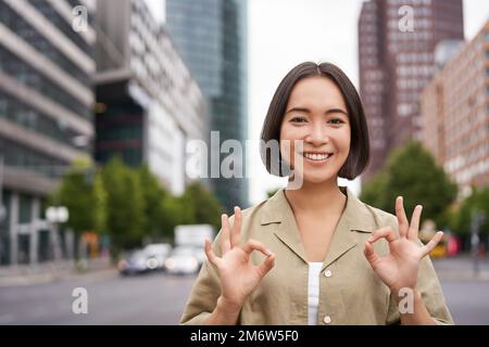 OK, Pas de problème. Souriante jeune femme asiatique montre ok geste, approuver smth, comme et d'accord, acceptant, debout sur la rue de cit Banque D'Images