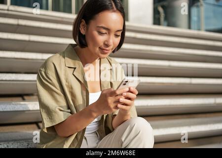 Les gens et la technologie. Belle femme asiatique souriante assise dans les escaliers de la ville, tenant le téléphone portable. Fille avec repose-smartphone Banque D'Images