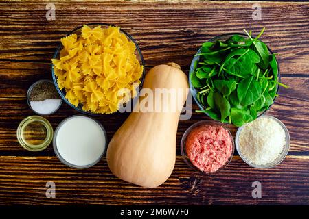 Pâtes de courge musquée crémeuse aux saucisses et aux épinards Ingrédients : pâtes farfalle, courge d'hiver et autres ingrédients crus sur une table en bois Banque D'Images