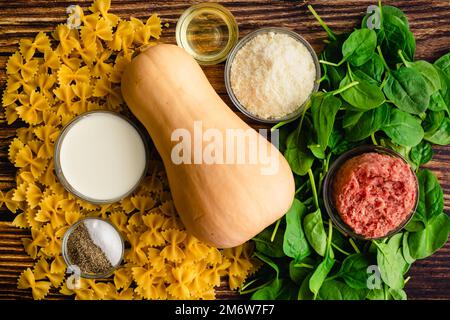 Pâtes de courge musquée crémeuse aux saucisses et aux épinards Ingrédients : pâtes farfalle, courge d'hiver et autres ingrédients crus sur une table en bois Banque D'Images