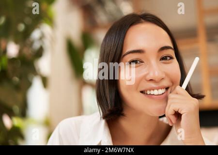 Portrait en gros plan de la jeune femme brune élégante, assis avec un stylo graphique et souriant, se détendre dans un café, écrire quelque chose, ma Banque D'Images