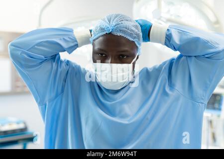 African american chirurgien masculin en blouse chirurgicale et chapeau nouant sur le masque dans le bloc opératoire Banque D'Images