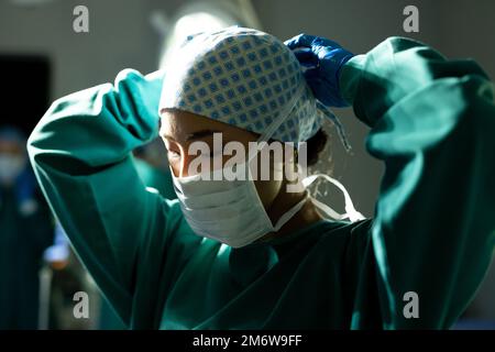 Femme asiatique chirurgien dans le chapeau chirurgical et robe nouant le masque dans le bloc opératoire Banque D'Images