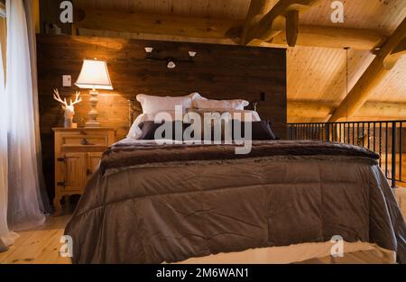 Lit queen size avec couvre-lit marron et table de nuit dans la chambre principale sur mezzanine à l'intérieur de maison en rondins de style cottage luxueux. Banque D'Images