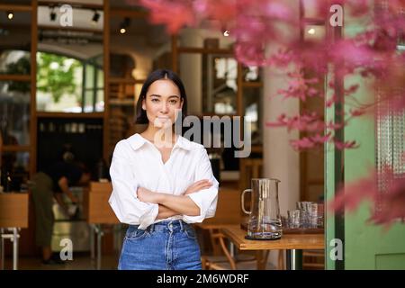 Portrait d'une jeune femme d'affaires dans son propre café, Manager debout près de l'entrée et vous invitant, posant dans une chemise blanche unie an Banque D'Images