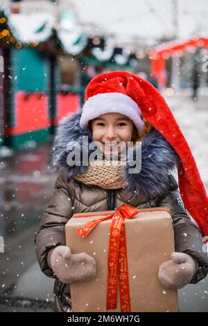 Portrait de fille joyeuse en chapeau de père Noël avec boîte cadeau pour Noël dans la rue de ville en hiver avec neige Banque D'Images