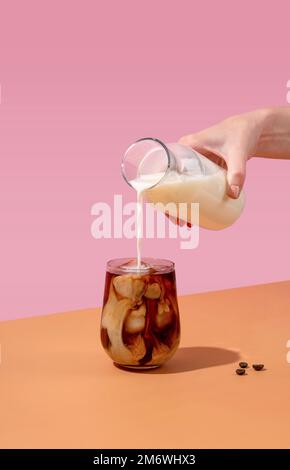 Du café glacé dans un grand verre avec de la crème versée et des grains de café. La main d'une femme verse de la crème dans un verre de café. Banque D'Images