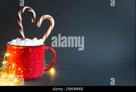 Tasse rouge avec guimauves et canne à sucre caramel en forme de coeur et lumières de guirlande sur fond noir. Noël Banque D'Images