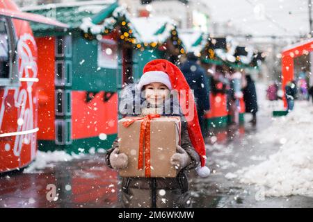 Portrait de fille joyeuse en chapeau de père Noël avec boîte cadeau pour Noël dans la rue de ville en hiver avec neige Banque D'Images