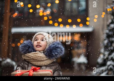Portrait d'une jeune fille joyeuse avec une boîte cadeau pour Noël dans une rue de ville en hiver avec de la neige sur un festival Banque D'Images