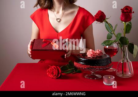 Femme en robe rouge main tenant une boîte cadeau rouge. Dîner de Saint-Valentin avec table, vin rouge, bougies et chocol Banque D'Images