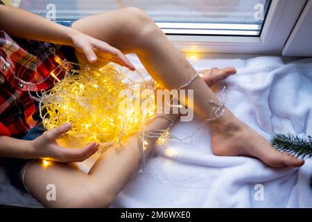 Une fille tient une guirlande entre ses mains, assise sur le rebord de la fenêtre et les fenêtres, ambiance festive, Noël, nouvel an Banque D'Images