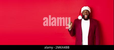 Concept de Noël, fête et vacances. Gai Black Guy dans le chapeau de santa pointant les doigts à gauche, montrant la bannière, debout amusé ag Banque D'Images