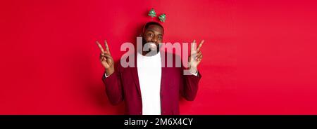 Joyeux Noël. Joyeux homme afro-américain barbu dans le bandeau de la fête, souriant et montrant des signes de paix, debout sur le bac rouge Banque D'Images