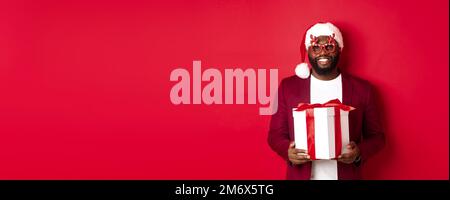 Noël. Beau homme afro-américain dans des lunettes de fête et chapeau de père noël tenant cadeau de nouvel an, apporter le présent dans la boîte et sourire, Banque D'Images