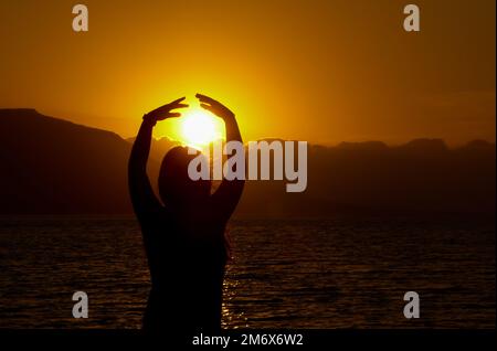 Silhouette de jeune femme à la mer en profitant du coucher du soleil. Lumière du soleil dans l'océan derrière les montagnes Banque D'Images