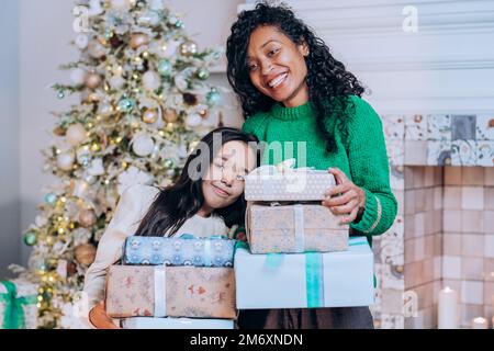 Joyeuse mère afro-américaine et fille brune avec de longs cheveux posent contre les décorations d'arbre de Noël tenant des cadeaux Banque D'Images