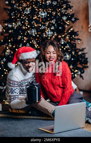 Homme noir et femme blanche dans les chandails de fête parlent de discuter par ordinateur portable et de montrer boîte cadeau contre l'arbre de Noël avec des ornements Banque D'Images