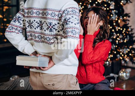 L'homme afro-américain en chandail à imprimé chaud surprend la brune mauriquement femme avec boîte de cadeau derrière dos contre l'arbre de Noël Banque D'Images