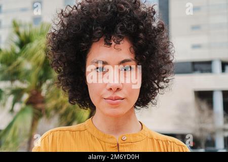 Gros plan d'une jeune femme hispanique avec des cheveux ondulés et bouclés regardant sérieusement l'appareil photo. Vue de face de la jolie dame latine brunette debout à l'extérieur. Photo de haute qualité Banque D'Images