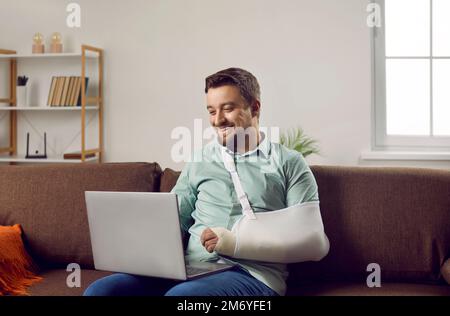 Un jeune homme heureux avec le bras dans un casting assis sur le canapé à la maison et communiquant sur un ordinateur portable. Banque D'Images