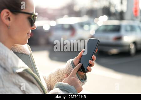 Femme utilisant son smartphone en touchant l'écran avec son doigt tout en se tenant sur le parking. Banque D'Images