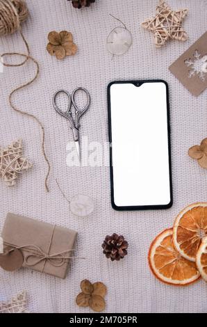 Concept d'emballage de cadeaux pour les vacances. Cadeau écologique artisanal, tranches d'orange séchées, fleurs, étiquette, ciseaux, cônes de sapin, corde sur fond rose. Haut du smartphone v Banque D'Images