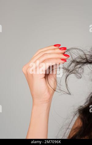 vue rognée de la femme brune avec manucure rouge tenant les cheveux endommagés isolés sur gris, image de stock Banque D'Images