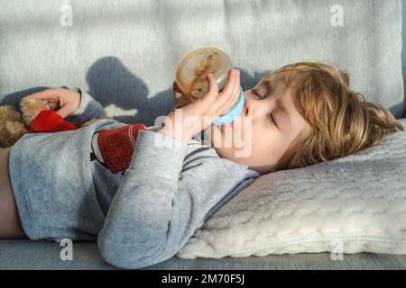 Portrait en gros plan de bébé garçon tenant et nourrissant le lait du biberon allongé sur le canapé. Un petit garçon mignon avec une bouteille de lait. Temps de sommeil. Banque D'Images