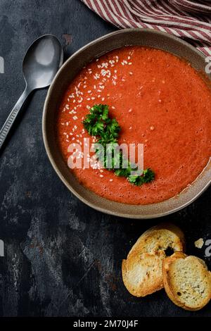 Gazpacho garni de persil dans une assiette à bords ondulés. Il y a du poivron rouge, de la tomate, de l'ail, des croûtons et une cuillère sur la table. Vue de dessus. Banque D'Images
