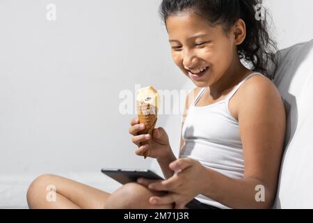 une petite fille brune heureuse et souriante mangeant une glace tout en jouant sur sa tablette ou son téléphone portable, assise sur le canapé dans son salon. Banque D'Images