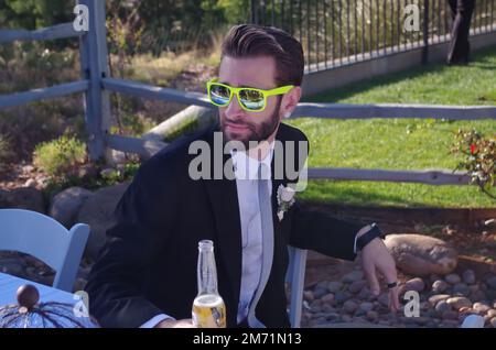 Jeune homme assistant à un événement formel portant des lunettes de soleil de style Ray-Ban aux couleurs vives, buvant une bière Banque D'Images