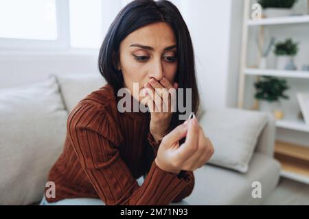 Une femme regarde malheureusement un anneau de mariage dans sa main, le divorce et la perte d'un partenaire, la rupture, la rupture de coeur malheureux assis à la maison sur le canapé Banque D'Images