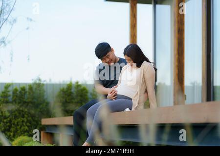 Femme enceinte asiatique avec son mari parlant ensemble en terrasse à la maison pour se détendre et respirer de l'air frais. Banque D'Images