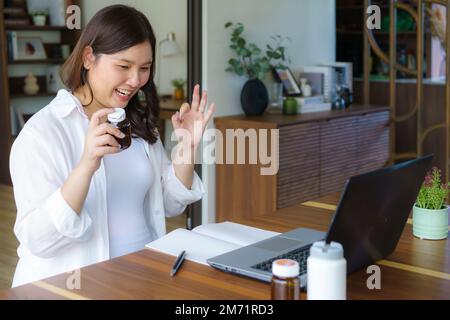 Femme enceinte asiatique tenant une bouteille de pilule communiquant avec le médecin sur le Web dans le salon à la maison. Télémédecine, grossesse, bébé attendu et santeca Banque D'Images